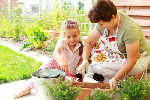 A woman and a young girl are engaged in gardening activities in a lush backyard garden during daylight hours. The woman, dressed casually in a green top and a colourful floral apron, is kneeling beside the girl and assisting with planting or potting soil into a rectangular planter box, which is situated on a paved patio area. The girl, wearing a light pink shirt and striped pink and white trousers, is focused on her task, holding a small gardening tool. In the foreground, there are potted plants with vibrant green foliage and small, delicate flowers, and a metal watering can is nearby. The background reveals a well-maintained garden with a variety of green hedges, shrubs, and trees, with a wooden fence bordering the space and some flower beds with lush plants. The overall scene suggests a bright, mild day, capturing a moment of outdoor gardening in a family-friendly garden setting near Whitechapel or the London area, reflecting the services of Gardening Services Whitechapel in outdoor maintenance and garden care.