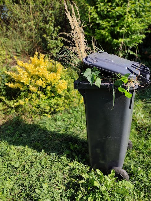 A black wheeled garden waste bin positioned on a grassy area in a garden, filled with various garden debris such as cut grasses, leaves, and plant stems. Behind the bin, there is a yellow flowering sh