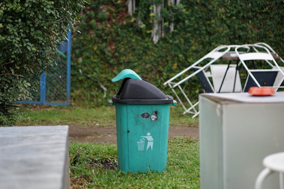 A black wheeled garden waste bin positioned on a grassy area in a garden, filled with various garden debris such as cut grasses, leaves, and plant stems. Behind the bin, there is a yellow flowering sh
